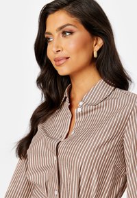 Camicia a righe marroni e bianche con colletto classico, patta dei bottoni e tessuto testurizzato. Modello accessoriaméntato con orecchini in oro.