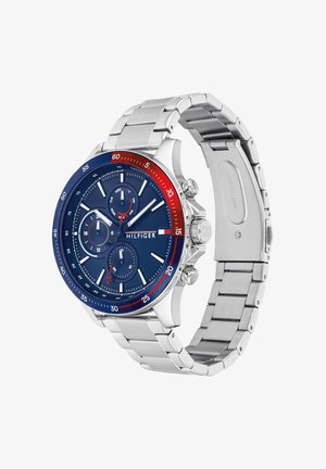 Zilveren horloge met metalen schakels, blauwe en rode rand, blauwe wijzerplaat, drie subwijzerplaten, witte markers en "Hilfiger"-logo op de wijzerplaat.