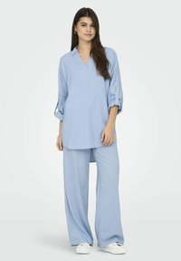 Chemise bleu clair, ample, à boutons et col en V, avec des manches retroussées, associée à un pantalon large assorti. Baskets blanches.