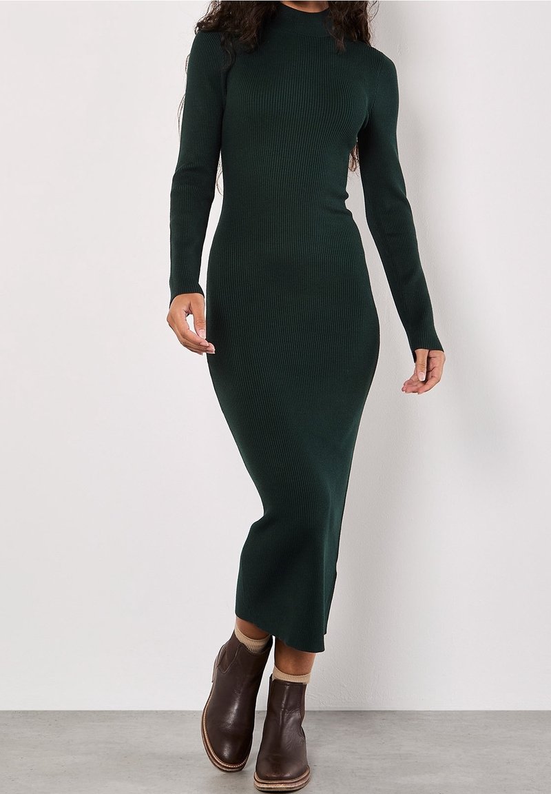 Femme portant une robe midi moulante à manches longues en maille côtelée vert foncé, associée à des bottines marron, marchant sur un sol gris.