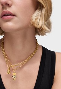 Un collier doré à plusieurs couches présente un design de chaîne épaisse avec deux pendentifs suspendus : un ours et un trèfle, tous deux dotés de détails intriqués.