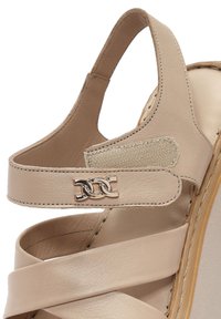 Sandalo in pelle beige con cinturini incrociati, una cintura posteriore alla caviglia e una placca decorativa in metallo con logo. Tessitura liscia e design arrotondato.