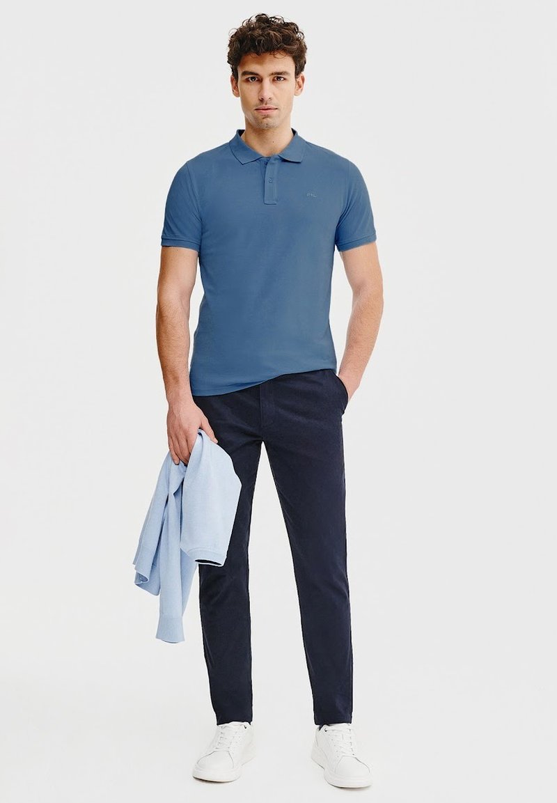 Jasnoniebieska koszulka polo z krótkim rękawem i kołnierzykiem, zestawiona z granatowymi spodniami i białymi sneakersami; trzyma jasnoniebieski sweter.