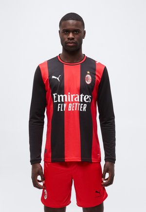 Mužský atlét oblečený v červeno-čiernom pruhovanom dresu AC Miláno s dlhým rukávom a červených šortkách, stojaci na jednoduchom bielom pozadí.