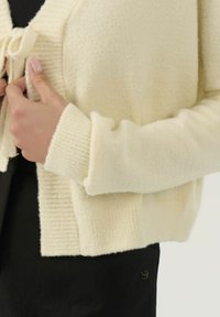 Persona che indossa un cardigan in maglia color crema, abbottonato su una maglietta nera, mostra un primo piano della manica e della mano che sistemano l'indumento.