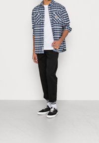 Blauwe en witte geruite button-up shirt met twee borstzakken over een wit T-shirt, gecombineerd met zwarte broek en zwarte sneakers met witte zolen.