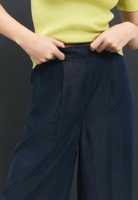 Aukštu juosmeniu tamsiai mėlyni platus kojos kelnių su klostėmis priekyje, pagaminti iš minkštos džinsinio audinio, derinami su siauro kampo limonado spalvos marškinėliais.