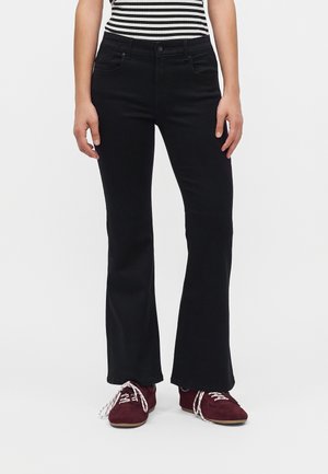 Person iført sorte flare jeans, en sort-hvidstribet top og bordeaux sko med hvide snørebånd, stående på en enkel baggrund.