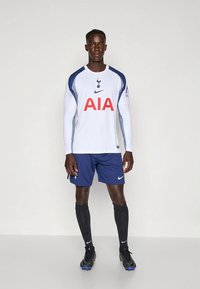 Fotbalový dres Tottenham Hotspur zahrnující bílý dlouhý rukáv s námořnickými akcenty a velkým červeným logem "AIA", doplněný o námořnické šortky.