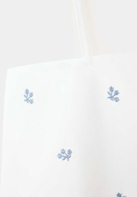 Sac fourre-tout en tissu blanc avec une fine bandoulière et une petite broderie florale bleue dispersée à la surface.