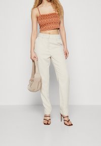 Haut orange court avec un motif ondulé, pantalon crème ajusté et sandales à talons brunes. Tient un sac à épaule en tissu beige.