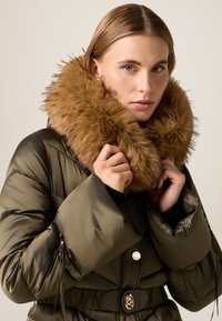 Chaqueta acolchada verde oliva con un gran cuello de piel sintética marrón, ceñida en la cintura con un cinturón negro, con detalle de botón dorado.