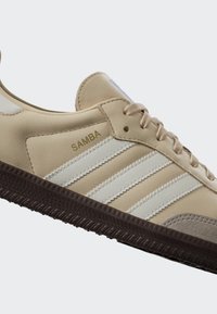 Béžová teniska Adidas Samba s bílými pruhy, hnědou texturovanou podrážkou, béžovými tkaničkami a nápisem "SAMBA" na straně.