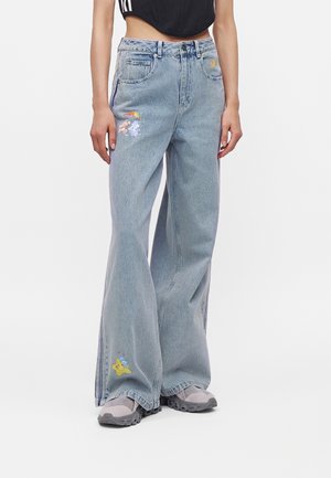 Persoon draagt lichtblauwe wijde jeans met kleine kleurrijke cartoonpatches en grijze sneakers, staand tegen een witte achtergrond.