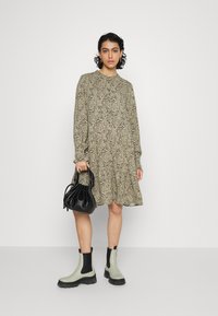 Vestido de manga longa com padrão floral em bege e oliva, com barra rodada. A modelo usa botas chelsea pretas e verdes e segura uma bolsa preta.