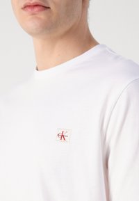 Weißes Baumwoll-T-Shirt mit rundem Ausschnitt und kurzen Ärmeln; verfügt über ein kleines Stoffetikett mit rotem "K"-Logo in der Nähe der Brust.