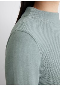 Helles grünes Sweatshirt mit strukturiertem Strickmuster und hohem, geripptem Halsausschnitt. Der Stoff wirkt weich und leicht dick.