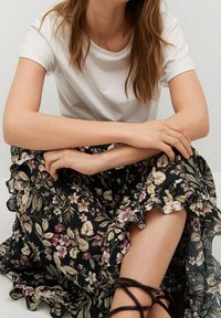 Haut blanc à manches courtes associé à une jupe noire à fleurs avec un ourlet à volants, présentant des fleurs roses et crème avec des feuilles vertes. Sandales noires à brides.