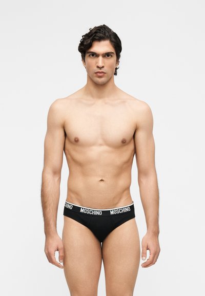 Άνδρας μοντέλο με σκουρόχρωμα μαλλιά που φοράει μαύρα εσώρουχα Moschino, στέκεται και κοιτάει προς τα εμπρός μπροστά σε απλό γκρι ανοιχτό φόντο.