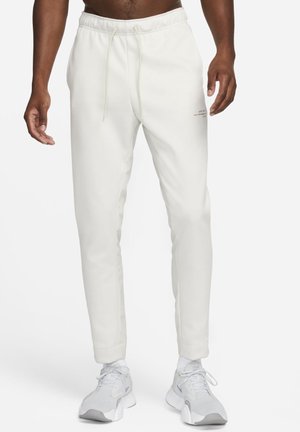 Man draagt off-white taps toelopende joggingbroek met trekkoord in de taille en witte Nike-sneakers, staand tegen een effen witte achtergrond.