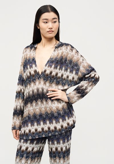 Γυναίκα με μακριά σκούρα μαλλιά που φοράει ταιριαστό πλεκτό τοπ και παντελόνι με βαθύ V και κυματιστό μπλε, μπεζ και λευκό μοτίβο, ποζάροντας με το χέρι στη μέση.
