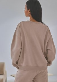 Übergroßer beiger Hoodie mit gerippten Bündchen und Saum, der eine lockere Passform und eine weiche Textur aufweist, von hinten gesehen.