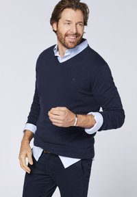 Marineblaues V-Ausschnitt-Pullover aus weichem Strickstoff, über einem hellblauen Hemd mit Kragen getragen. Verfügt über ribbelige Bündchen und ein kleines Logo auf der Brust.