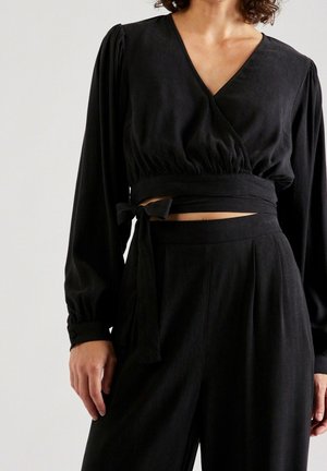 Kobieta ubrana w czarny, długi rękaw, krótki top z kopertowym zapięciem wiązanym w talii oraz w wysokim stanie czarne spodnie na tle jednolitego tła.