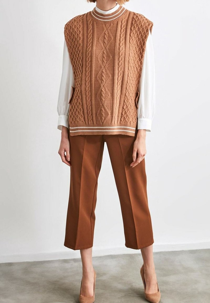 Personne portant un pull sans manches marron en maille torsadée sur un chemisier blanc, un pantalon marron raccourci et des talons hauts beige, debout sur un sol gris.