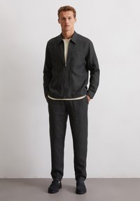 Dark gray, zippered jacket and matching trousers made of linen. Features a front pocket, relaxed fit, and worn with a beige shirt underneath.