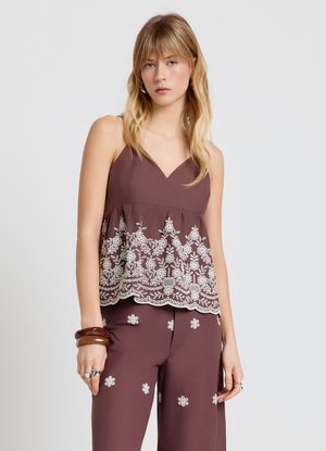 Jeune femme portant un débardeur marron avec broderie florale blanche et un pantalon marron assorti avec des fleurs blanches, accessoirisée de bracelets et de bagues.