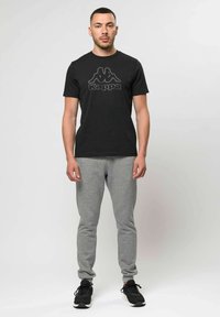 Mand stående med front mod kamera, iført sort Kappa T-shirt, grå joggingbukser og sorte sneakers mod en ensfarvet lys baggrund.