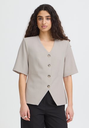 Femme aux longs cheveux bouclés portant un chemisier beige clair à manches courtes avec quatre boutons et un pantalon noir devant un fond uni.