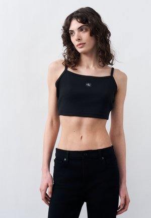 Giovane donna con capelli ricci che indossa un top nero corto e pantaloni neri, in piedi contro uno sfondo chiaro uniforme.