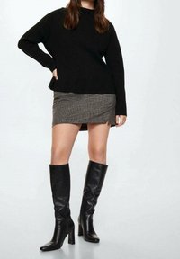 Schwarzer Strickpullover, Hahnentrittmuster-Minirock und kniefeine Lederstiefel mit Blockabsatz. Der Pullover hat eine lockere Passform und einen hohen Kragen.