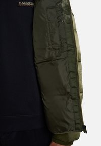 Manica di giacca imbottita verde oliva con polsini a costine, tasca con zip e tessuto blu scuro a contrasto sotto. Texture liscia e materiale sintetico.