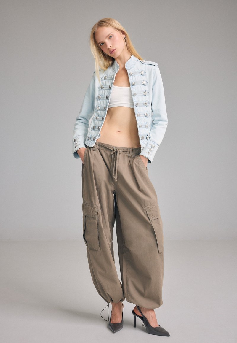 Jeune femme portant une veste bleu clair style militaire, un top court blanc, un pantalon cargo marron oversize et des chaussures à talons noires, debout devant un fond uni.