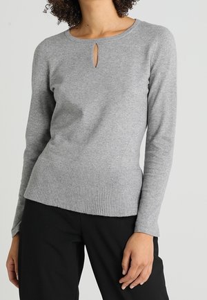 Kvinde iført en tætsiddende grå langærmet sweater med en lille nøglehulskæring og ribbet kant, kombineret med sorte bukser.