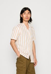 Vartalosetä, kaulushihnalla varustettu lyhythihainen paita, jossa on pystysuuntaiset beige- ja valkoviivat. Kangas näyttää kevyeltä ja rento istuvuus, yhdistettynä oliivinvihreisiin cargo-shortseihin.