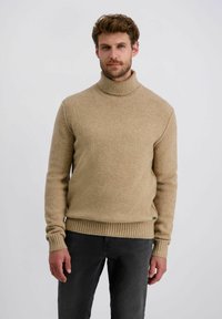 Beige Rollkragenpullover aus strukturiertem Strickstoff, mit gerippten Bündchen und Saum, getragen über dunkelgrauen Hosen.