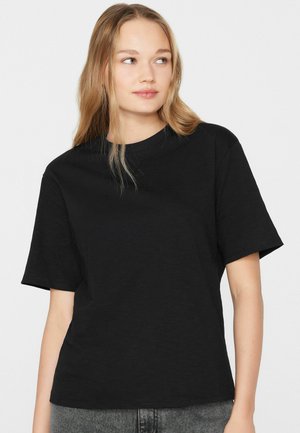 Junge Frau mit langen blonden Haaren, die ein schlichtes schwarzes T-Shirt mit kurzen Ärmeln und graue Hosen trägt, seitlich blickend vor weißem Hintergrund.