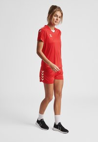 Ensemble de t-shirt et de shorts de sport rouges avec des accents blancs et des motifs en chevrons sur les shorts. Le modèle porte des baskets noires et des chaussettes blanches.