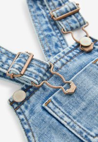 Jeanslatzhosen aus hellblauem Stoff mit kupferfarbenen Beschlägen, verstellbaren Trägern und einer Vorderseite mit sichtbaren Nähdetails.
