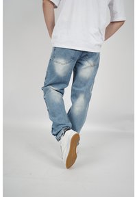 Jeans de mezclilla de lavado claro con un ajuste relajado, que presentan detalles ligeramente desgastados y dos bolsillos traseros, combinados con zapatillas blancas.