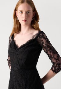 Robe en dentelle noire avec des manches trois-quarts, décolleté en V et taille cintrée. Présente des motifs en dentelle complexes et un tissu texturé.