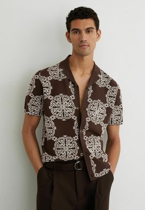 Mladý muž s krátkymi kučeravými vlasmi, oblečený v hnedej krátkorukávej košeli na gombíky s bielymi zložitými kruhovými vzormi, ruky v tmavohnedých nohaviciach vo vreckách.