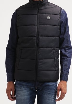 Homme portant une veste sans manches matelassée noire par-dessus une chemise sombre à manches longues et un jean bleu, debout avec les bras détendus le long du corps.
