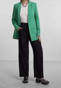Femme portant un blazer vert, une chemise blanche, un pantalon large noir, un collier en chaîne épais et des mocassins à talons métalliques noirs, se tenant devant un fond uni.