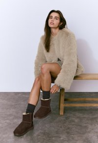 Botines marrones de ante con forro de piel, combinados con calcetines oscuros, un suéter beige peludo y una falda a cuadros, sentada en un banco de madera.