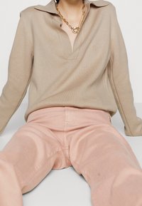 Persoon zittend in een beige lange mouw top met kraag en een gouden kettinghanger, gecombineerd met lichtroze broek.